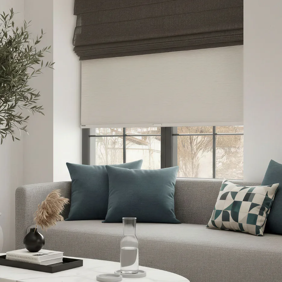 Contemporary living room featuring a gray upholstered sofa positioned beneath large windows with white and dark roller shades, styled with teal and patterned pillows, white coffee table with water ...