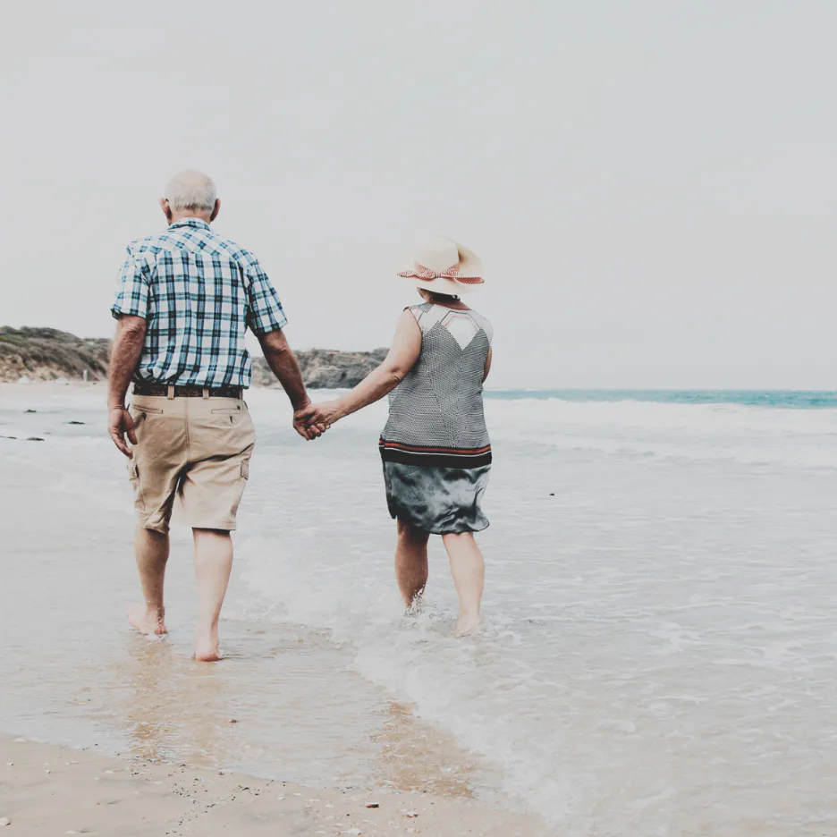 An older man and woman hold hands while walking barefoot through shallow ocean water on a sandy beach, wearing casual summer clothing and wide-brimmed hats under a clear sky.