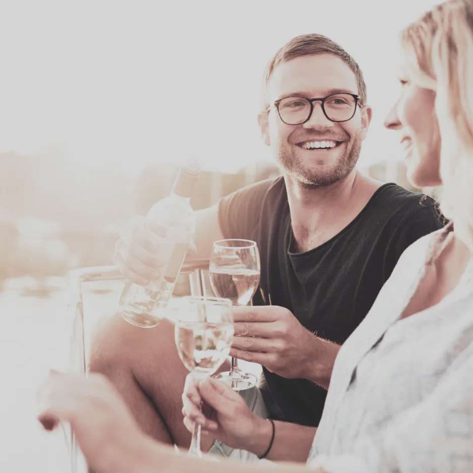 Smiling man wearing glasses and black shirt toasts with water glasses alongside a woman in professional setting, warm natural lighting