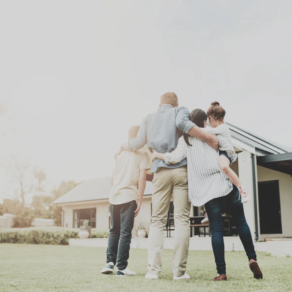 Four people standing on a grassy lawn with arms around each other in a group hug, facing a contemporary white house with a sloped roof in the background under an overcast sky.