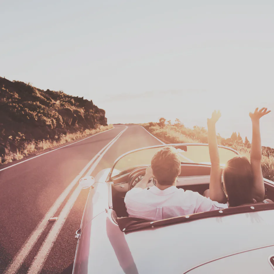 Two people in a white convertible with arms raised, driving down a desert highway with mountains and golden sunlight, capturing carefree adventure and freedom.