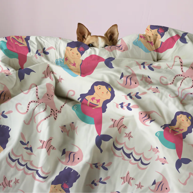 Tan dog peeking out from white kids bedding featuring illustrated mermaids in pink and navy, colorful fish, and ocean-themed patterns with decorative foliage elements