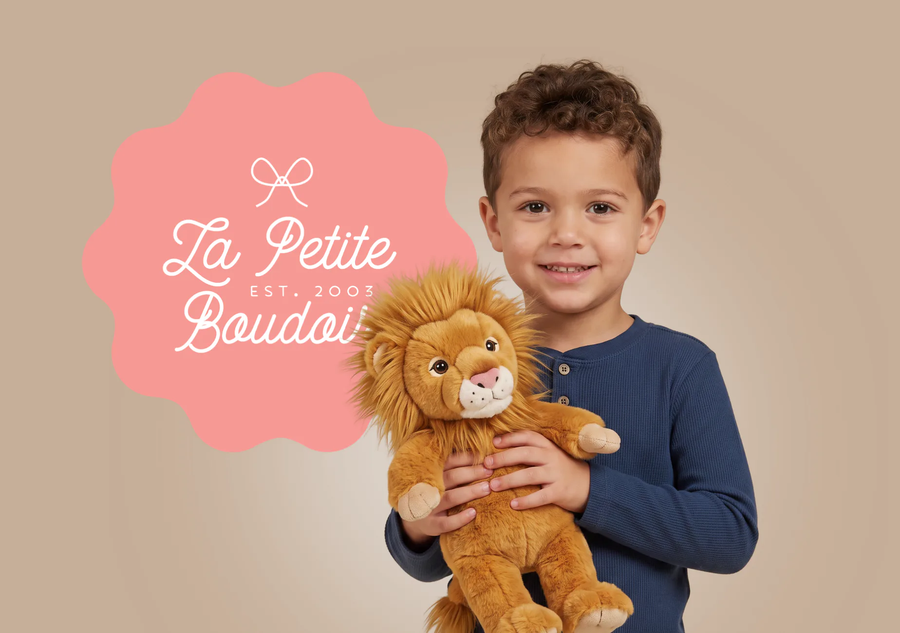 Young boy in navy shirt holding a plush golden lion toy, smiling at camera with pink branded badge behind him displaying 'La Petite Boudoir EST. 2003' logo