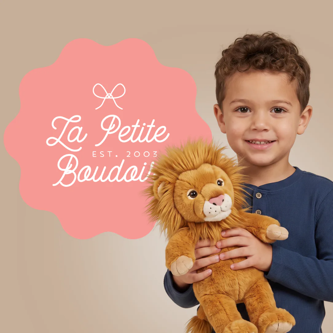 Young boy in navy shirt smiling while holding a golden plush lion toy, with pink scalloped badge displaying 'La Petite Boudoir EST. 2003' logo on neutral beige background.