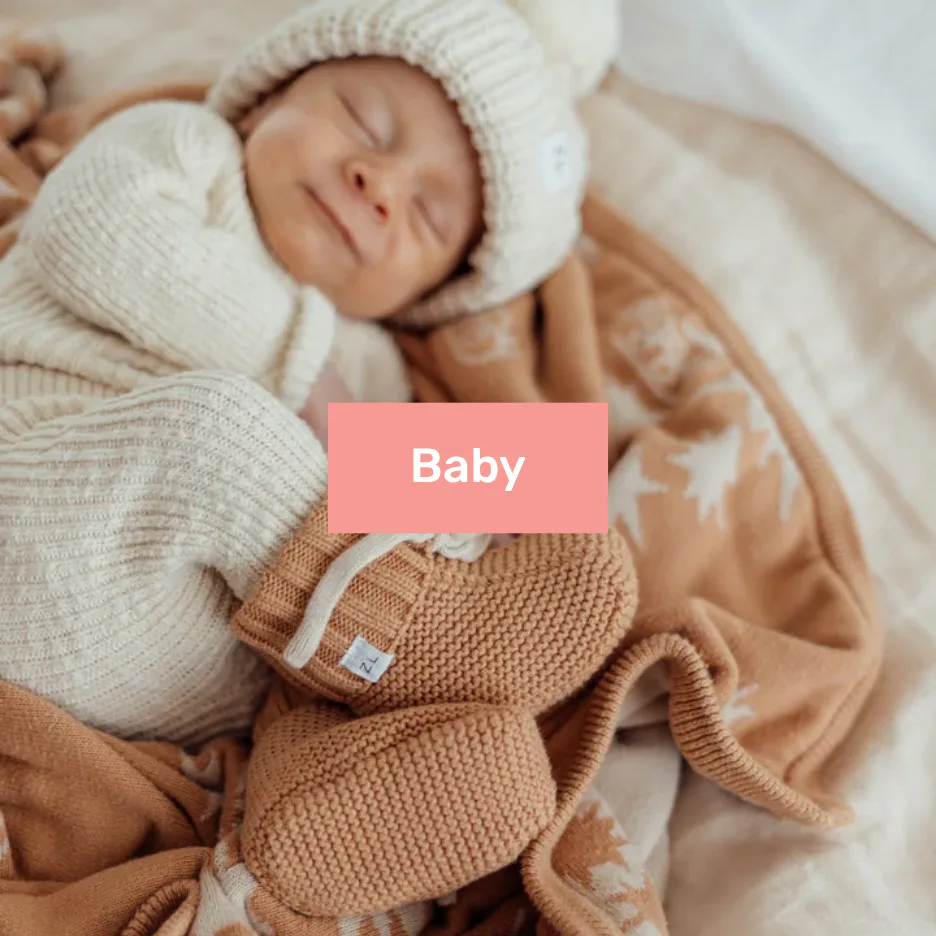 Sleeping newborn infant wearing a cream knitted hat and tan knit sweater, wrapped in soft neutral-toned blankets and bedding
