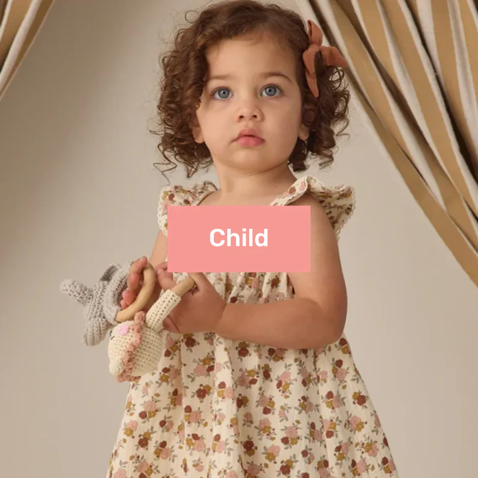 Toddler with curly brown hair and blue eyes wearing a cream floral print dress with a coral bow, holding a crocheted cream rabbit toy against a beige curtained background.